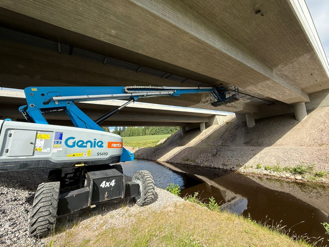 Henkilönostin puron rannalla betonisillan alla, työkori nostettuna sillan kannen alapintaan.